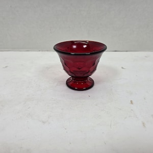 May include: A ruby red glass goblet with a textured, geometric pattern on the bowl. The goblet has a rounded base and a flared rim. The glass appears to be vintage or antique.