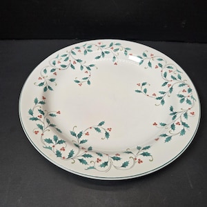 May include: White ceramic serving plate with a green and red holly berry design around the rim. The plate has a scalloped edge and a dark green border. A festive piece for holiday entertaining.
