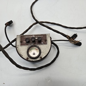 Könnte beinhalten: Ein Vintage-Plattenspieler von Webster Chicago mit einem runden Zifferblatt und drei Knöpfen mit den Beschriftungen "Radio", "Aufnahme" und "Wiedergabe". Das Gerät ist mit einem schwarzen Kabel mit mehreren Steckern verbunden.