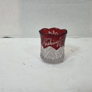 Puede incluir: Un pequeño vaso de chupito de recuerdo con una sección superior de color rojo intenso con el texto en blanco Washington D.C. y una base de vidrio prensado transparente y facetado con un patrón decorativo de corazones y diamantes.