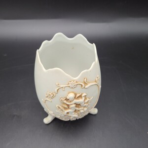 Puede incluir: Maceta de cer&aacute;mica blanca en forma de huevo con un dise&ntilde;o en relieve de querubines y motivos florales. La maceta tiene tres patas y es perfecta para exhibir plantas o flores peque&ntilde;as.