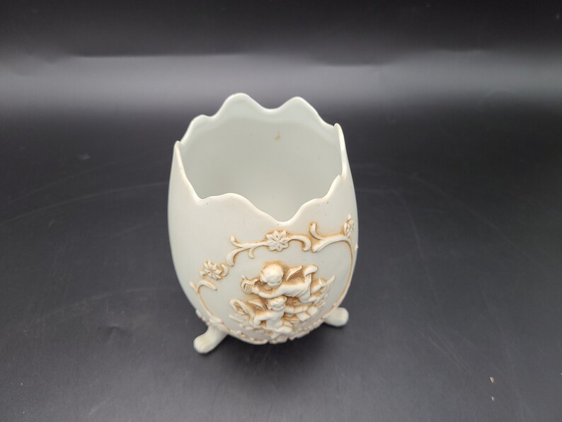Puede incluir: Maceta de cer&aacute;mica blanca en forma de huevo con un dise&ntilde;o en relieve de querubines y motivos florales. La maceta tiene tres patas y es perfecta para exhibir plantas o flores peque&ntilde;as.
