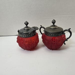 May include: A red glass creamer and sugar bowl set with silver-toned lids and handles. The red glass has a textured, frosted appearance. The sugar bowl has two handles. The creamer has a spout.
