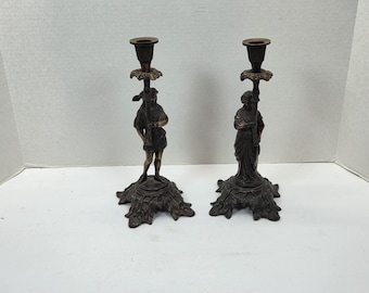 May include: Pair of antique bronze candle holders. Each holder features a detailed figure, one in a tunic and the other in a flowing gown, atop a decorative base. The candle cups are a contrasting bronze color.
