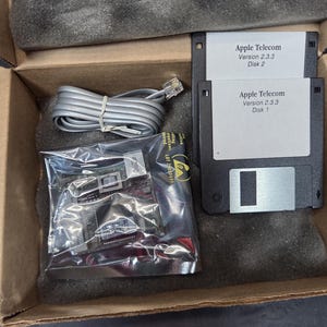 May include: An open cardboard box containing computer hardware. Inside are two 3.5-inch floppy disks labeled "Apple Telecom Version 2.3.3 Disk 1" and "Disk 2". Also included is a network cable and a circuit board in a plastic bag.