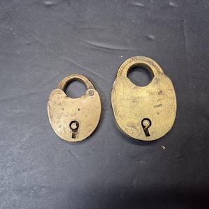 Può includere: Due antichi lucchetti in ottone di diverse dimensioni. I lucchetti hanno una forma ovale con una serratura e un manico arrotondato. L'ottone ha un aspetto usurato e invecchiato, suggerendo che si tratta di oggetti d'epoca.