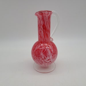 May include: A red and white glass pitcher with a clear handle and a small spout. The pitcher has a round base and a tall, cylindrical body. The glass has a speckled pattern.