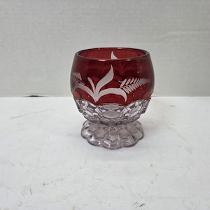 Puede incluir: Un pequeño cuenco de cristal redondo con una sección superior de color rojo intenso con patrones de hojas de helecho grabados en blanco. La parte inferior del cuenco y la base circular son de cristal transparente con un patrón geométrico de panal.