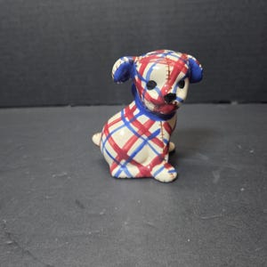 May include: A ceramic dog figurine with a plaid pattern in red and blue on a cream background. The dog has blue ears and a blue collar. The figurine is sitting down and has a black nose.