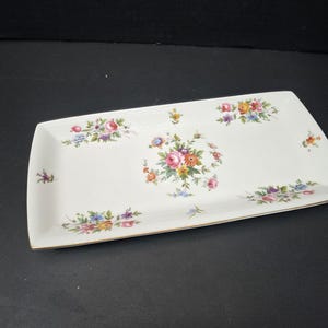 Könnte beinhalten: Rechteckige weiße Keramik-Servierplatte mit abgerundeten Ecken. Die Platte ist mit Blumensträußen in Rosa, Gelb, Blau und Lila verziert. Der Rand hat eine goldene Zierleiste.