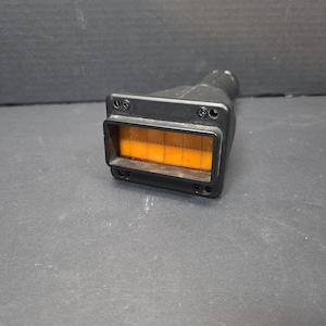 Puede incluir: Un dispositivo rectangular negro con una ventana teñida de naranja y con patrón de cuadrícula. El dispositivo tiene una extensión cilíndrica y está asegurado con tornillos. El objeto está sobre una superficie gris oscuro.