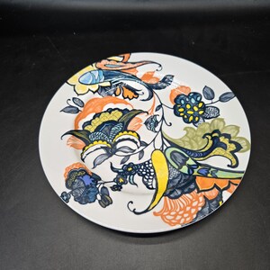 May include: A white ceramic dinner plate with a colorful floral design. The design features orange, yellow, blue, green, and black flowers and leaves.