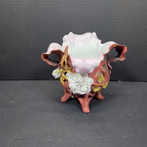 Op de afbeelding: Een decoratieve keramische vaas met een bloemenontwerp. De vaas is voornamelijk roodbruin met roze en witte accenten. Het heeft een bloemachtige vorm, drie kleine voetjes en een witte bloem aan de zijkant.