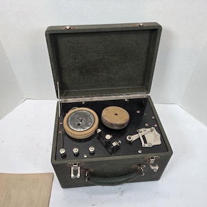 May include: A vintage radio receiver housed in a dark green case with a hinged lid. The interior has a black panel with dials, knobs, and components. A handle and latches are visible on the front.