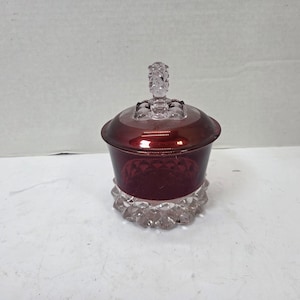 May include: A small, round glass candy dish featuring a deep red, smooth upper section and a clear, faceted, diamond-patterned base. The matching red glass lid has a clear, textured, pineapple-shaped finial handle on top.