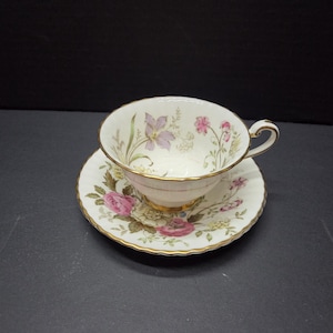 Könnte beinhalten: Ein Vintage-Tee-Set. Die weiße Porzellantasse und Untertasse sind mit rosa und lila Blumenmustern und goldenen Verzierungen verziert. Die Tasse hat einen geschwungenen Henkel.