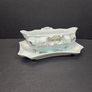 May include: A white porcelain gravy boat with a matching saucer. The boat has a rectangular shape with ornate handles and a decorative design in shades of blue, brown, and white. The saucer has a scalloped edge.