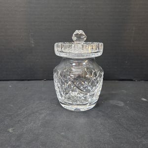 May include: A clear, cut-glass jar with a lid. The jar has a rounded body and a lid with a faceted knob. The glass is intricately cut with a repeating pattern. The jar is likely used for storing small items.