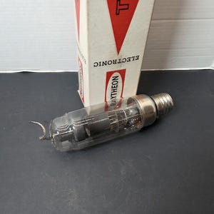Puede incluir: Un tubo de vacío electrónico Raytheon vintage con una carcasa de vidrio transparente y una base de metal. El tubo está junto a su caja original, que tiene un triángulo rojo y la palabra "ELECTRONIC" impresa.