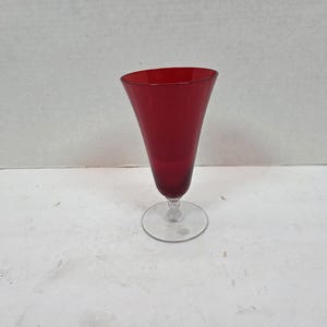 May include: A tall, ruby-red glass with a flared top and a clear, round base. The stem is short and features a decorative, textured design. The glass is likely used for serving beverages.