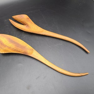 May include: A set of two wooden salad servers with a unique curved design. The servers are made of natural wood and have a warm brown colour.