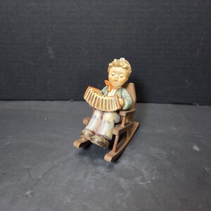 May include: A figurine of a young person playing an accordion while seated in a wooden rocking chair. The figurine has a detailed painted design with a green jacket, orange bow tie, and tan pants. The accordion is tan and brown.