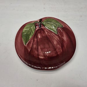May include: A ceramic plate shaped like a red apple with green leaves. The plate has a glossy finish and is designed to resemble the fruit. The leaves are a vibrant green, and the apple is a deep red.