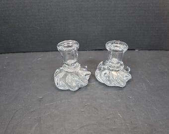 May include: Two clear glass candle holders. Each has a round top with a small opening for a candle and a base with a swirling, petal-like design. The candle holders are on a dark surface.