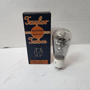 Pode incluir: Uma válvula de vácuo vintage e sua caixa original. A caixa azul marinho apresenta letras douradas que dizem "Taylor Custom Built Tubes". O tubo transparente tem componentes internos e uma base branca.