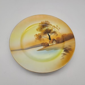 May include: A vintage ceramic plate featuring a hand-painted scene of a small house by a lake with a tree in the background. The plate has a yellow and orange background with brown accents.