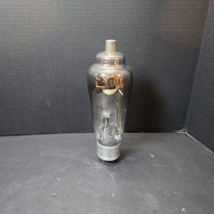 May include: A vintage vacuum tube with a clear glass body and a silver metal base and top. The tube has internal components visible through the glass. The base has white text. The tube is standing upright against a dark background.