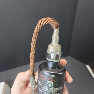 May include: A vintage Westinghouse Ignitron tube featuring a black cylindrical body with white printed text and a logo. A thick, braided copper wire cable extends from the top metal terminal, curving downward to a secondary metal connector point.