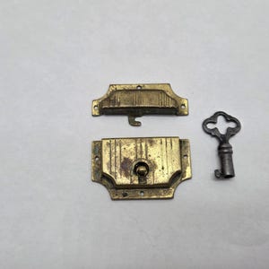 May include: An antique brass-coloured lock set with a key. The set includes the lock's two parts and a decorative key. The lock has a rectangular shape with a central keyhole and mounting holes. The key is dark grey.