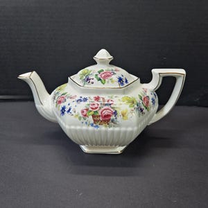 May include: A white porcelain teapot with a hexagonal body and a matching lid. The teapot features a colourful floral pattern depicting pink roses, blue cornflowers, and green leaves, with a small basket motif and gold trim on the edges.