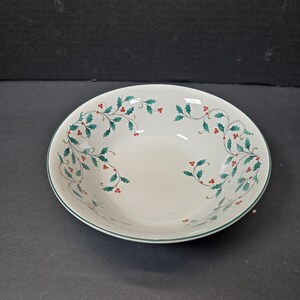 May include: A white ceramic bowl with a green rim and a decorative holly and berry pattern. The bowl has a shallow, rounded shape, suitable for serving soups or salads. The holly design features green leaves and red berries.