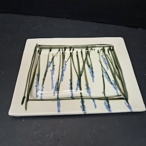May include: A rectangular, off-white ceramic plate with a hand-painted design. The design features vertical, dark green and blue brushstrokes, resembling reeds or grass. The plate has a slightly raised edge and a glossy finish.