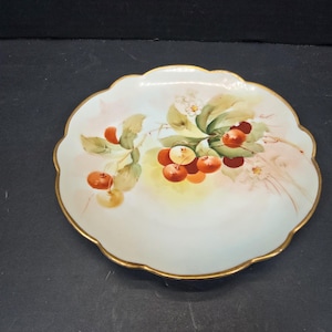 May include: A white, scalloped-edge plate with a gold rim. The plate features a hand-painted design of cherries with green leaves and small white flowers. The cherries are red and yellow, and the leaves are green.