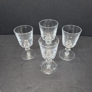 May include: Four clear, stemmed glasses with etched vertical lines and a round base are arranged on a dark surface. The glasses are of varying heights, suggesting a set of drinkware for different beverages.