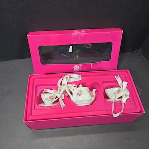 May include: A miniature porcelain tea set featuring a teapot, two teacups, and two saucers, all decorated with pink rose patterns and small dots. The set is presented in a bright pink rectangular gift box with a clear window and white ribbons.