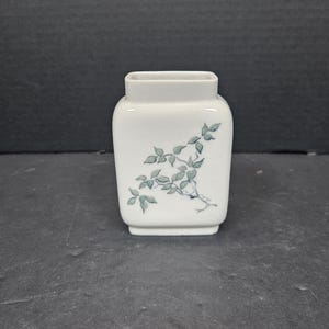 Puede incluir: Un pequeño jarrón de cerámica blanca de forma cuadrada. El jarrón presenta un delicado diseño de hojas y vides verdes en un lado. El jarrón es adecuado para pequeños arreglos florales o fines decorativos.