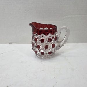 May include: A small glass creamer pitcher featuring a clear base and a deep red rim. The body is decorated with a raised, faceted diamond pattern, with the red color accenting the center of each diamond shape. It includes a curved handle and spout.