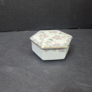 May include: A hexagonal porcelain trinket box with a cream-colored base and a lid featuring a delicate floral pattern of pink and blue flowers with green leaves. The edges of the lid are trimmed with a thin gold line, and the box is white.