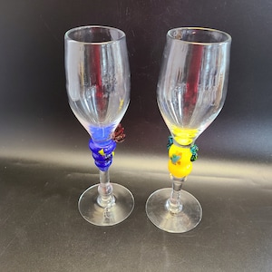 May include: Two clear glass champagne flutes with colourful glass beads around the stems. One flute has blue and yellow beads, the other has yellow and green beads.