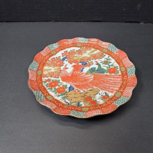 May include: A decorative ceramic plate with a scalloped edge. The plate features a central design of a red bird with a long tail, surrounded by flowers and foliage in orange, green, and gold. The rim has a patterned border.