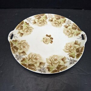 May include: Oval serving platter with a white background and a floral rose design in shades of beige, brown, and green. The platter has two handles and is suitable for serving food.