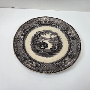 May include: A round, antique plate with a black and cream color scheme. The plate features a detailed landscape scene in the center, surrounded by ornate scrollwork and floral patterns. The plate's edge has a scalloped design.
