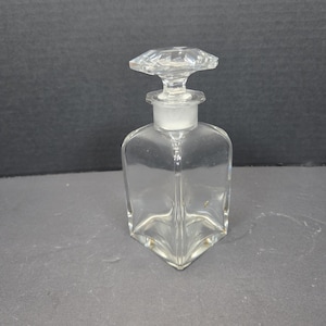 May include: A clear glass perfume bottle with a square base and a faceted, diamond-shaped stopper. The bottle has a white stopper and a clear, transparent body. The design is simple and elegant, suitable for displaying perfumes or colognes.