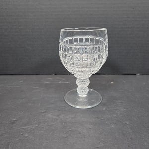 May include: A clear, cut-glass wine glass with a textured, grid-like pattern on the bowl. The stem features small, round details, and the base is a simple, circular design. The glass is empty and sits on a dark surface.