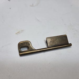 May include: A vintage metal key with a rectangular head and a curved handle with the text "DETEX". The key is a tarnished brass colour and appears to be old. The key is lying on a white surface.