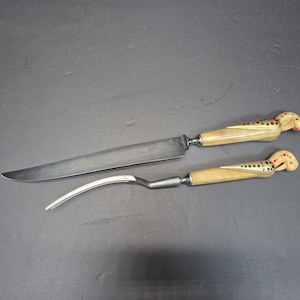 May include: A carving knife and fork set. The knife has a long, silver blade and a handle with a light brown finish and decorative accents. The fork has a curved silver tine and a matching handle. The handles have a bird-like design.